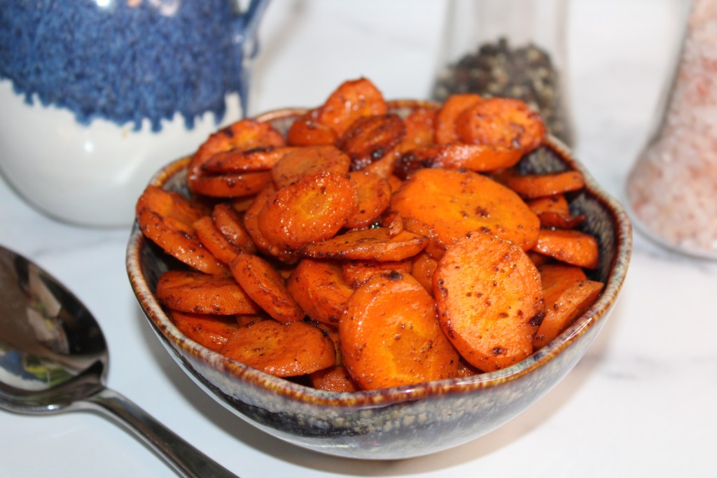 Bowl of roasted carrots with a golden-brown color, garnished with spices, placed on a marble countertop.
