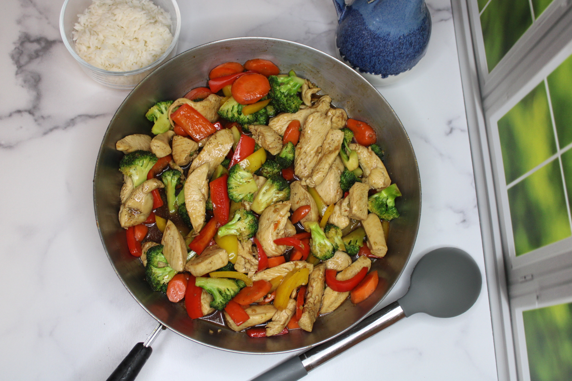 A stainless steel pan filled with stir-fried chicken strips and colorful vegetables, including red and yellow bell peppers, broccoli, and carrots, alongside a bowl of fluffy white rice.